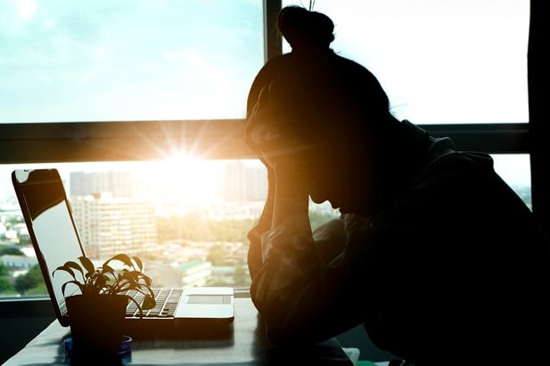 woman silhouetted at desk in front of laptop, hands to forehead