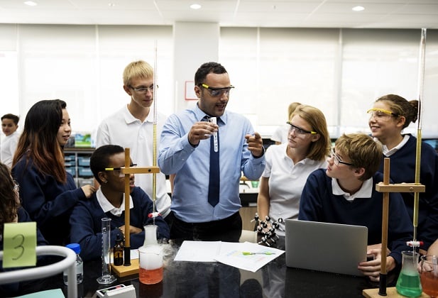 teacher wearing shirt and tie surrounded by students as he explains a lesson
