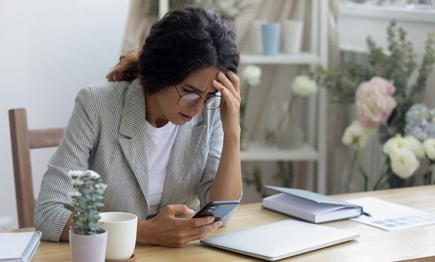 business woman at home desk looking at phone with great dismay and hand to forehead