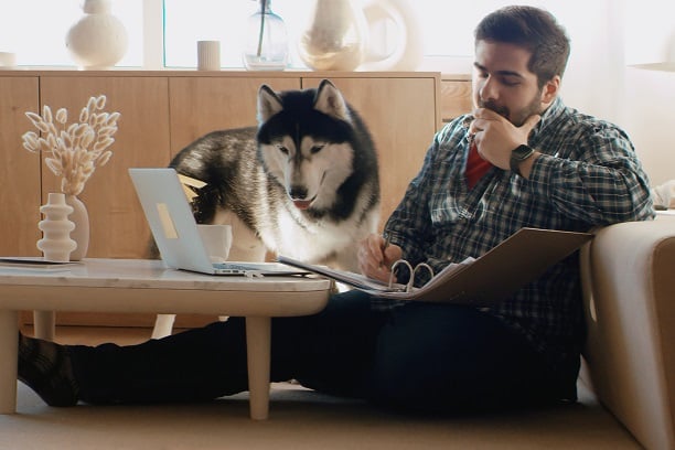 Man sitting beside sofa with laptop and his dog beside him and a notebook on his lap
