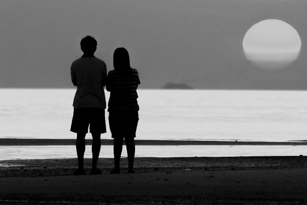 man and woman silhouetted as they stand by a lake watching rising moon