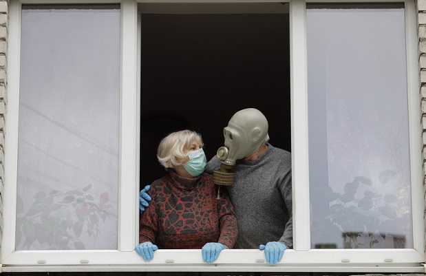 Two masked senior citizens, one in a big mask, stand at open window hugging