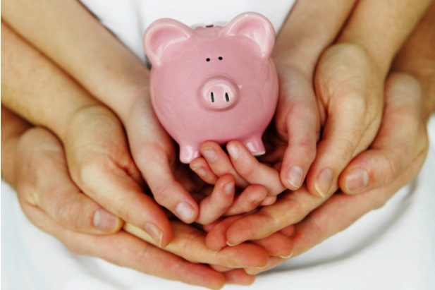 two pairs of adult hands and one pair of child hands nestled together and holding a pink piggy bank