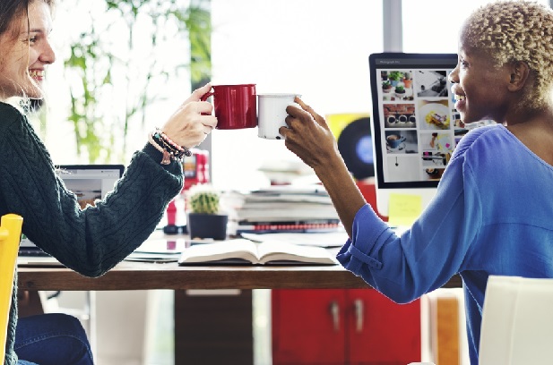 Two women at desks toasting each other with coffee mugs